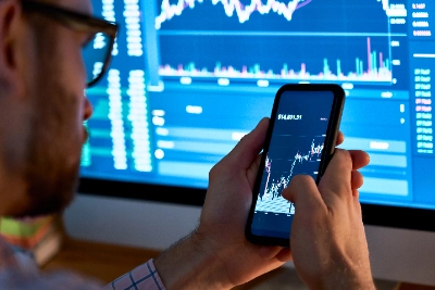 Un hombre con gafas analiza gráficos financieros en su smartphone mientras sostiene el dispositivo con ambas manos, justo enfrente de un gran monitor que despliega pantallas de datos y estadísticas del mercado bursátil en tonos azules.