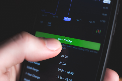 Un primer plano muestra el pulgar de una persona presionando un botón verde brillante que dice "Start Trading" en la pantalla de un smartphone, la cual despliega un gráfico de acciones y datos numéricos sobre un fondo oscuro.