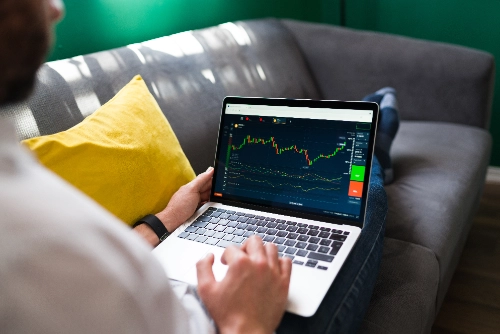 Un joven aparece relajado en un sofá gris con un cojín amarillo mientras observa un gráfico financiero de velas (trading) en la pantalla de su laptop