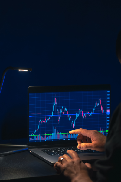Una persona analiza un gráfico de velas en un portátil bajo la luz de una lámpara de escritorio. La mano del usuario señala una tendencia en la pantalla, destacando el seguimiento activo de los mercados financieros.