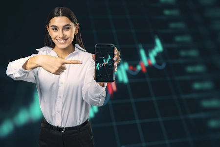 una mujer con camisa blanca sonríe mientras señala un gráfico financiero en su celular frente a un fondo digital oscuro.