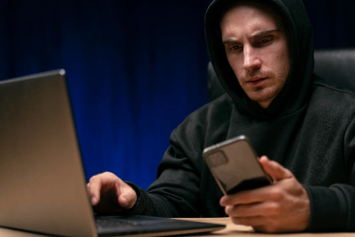 Un hombre con capucha negra mira con expresión preocupada su teléfono móvil mientras mantiene su otra mano sobre el teclado de una computadora portátil.