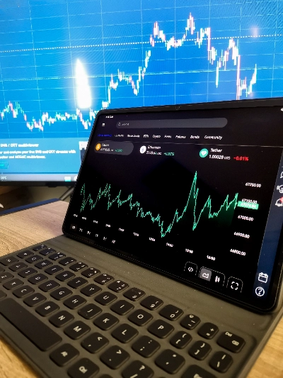 Una tableta con teclado acoplado muestra un gráfico financiero de criptomonedas con una línea verde sobre fondo negro, detallando activos como Bitcoin, Ethereum y Tether.