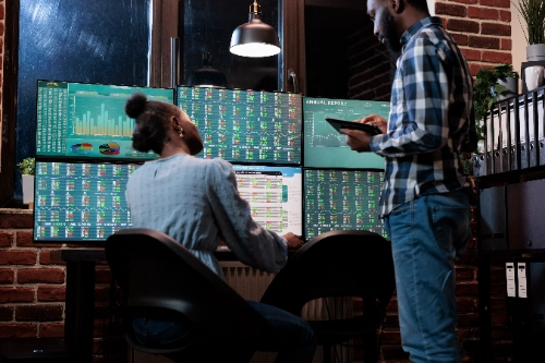 Dos analistas financieros trabajan de noche en una oficina frente a una estación de múltiples monitores que muestran gráficos de barras y tablas de datos del mercado de valores.