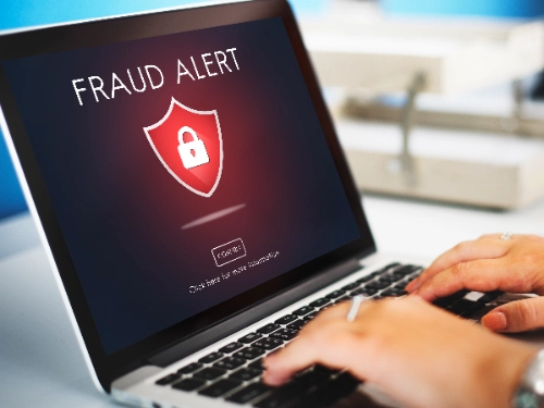 Un primer plano de unas manos utilizando una computadora portátil, en cuya pantalla se muestra un aviso de advertencia rojo que dice "FRAUD ALERT" junto al icono de un escudo con un candado.