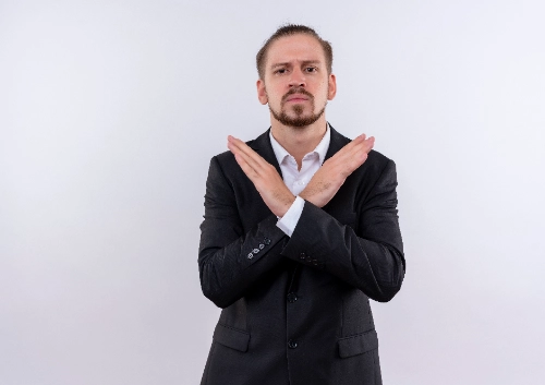 Un hombre de negocios con traje negro y expresión seria hace un gesto de rechazo o de detenerse cruzando los brazos en forma de "X" sobre un fondo blanco.