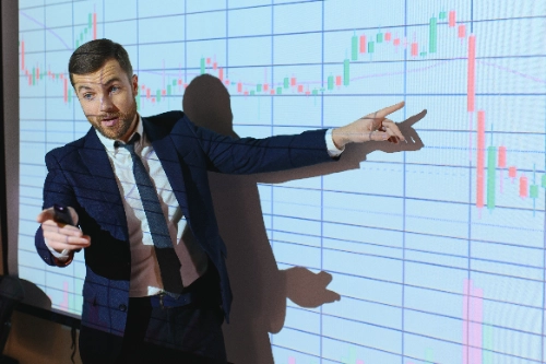 se muestra a un hombre en traje dando una presentación, apuntando con su mano hacia una gran pantalla proyectada que muestra un gráfico financiero de velas japonesas.