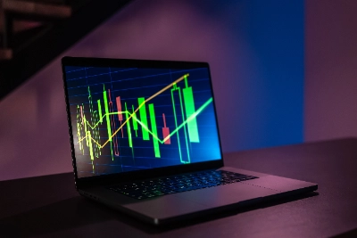 Un ordenador portátil abierto sobre una mesa oscura muestra en su pantalla un gráfico de velas japonesas verdes y rojas con líneas de tendencia ascendentes