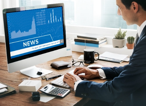 se ve de perfil a un hombre en traje trabajando frente a un monitor que muestra noticias financieras y gráficos azules