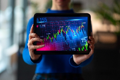 se muestra en primer plano una tableta sostenida con ambas manos por una persona con suéter azul. La pantalla del dispositivo despliega un gráfico financiero detallado con líneas de tendencia y velas japonesas en tonos verdes, rojos y morados.