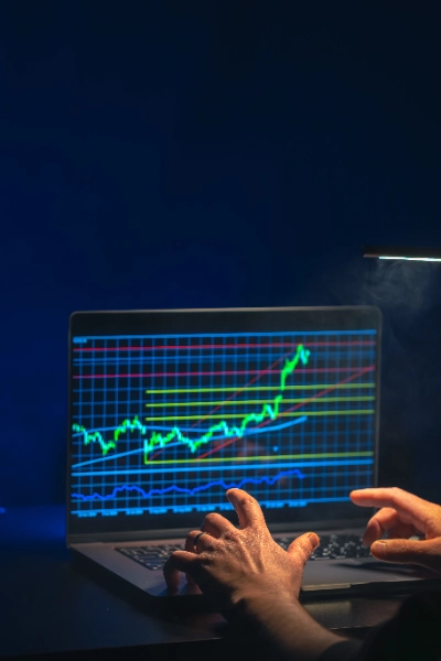 Unas manos escriben en el teclado de un ordenador portátil. La pantalla muestra un gráfico de trading detallado con cuadrícula azul, líneas de soporte amarillas y rojas, y una gráfica de velas verdes en tendencia ascendente.