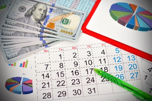 plano cenital que muestra un calendario mensual con un bolígrafo verde encima, rodeado de fajos de billetes de 100 dólares y varios gráficos estadísticos de pastel y de barras.