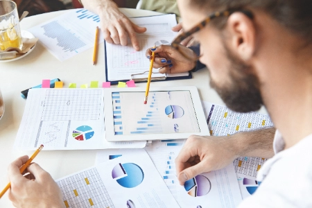 plano cenital de varios compañeros de trabajo analizando gráficos estadísticos, reportes impresos y una tableta digital sobre una mesa