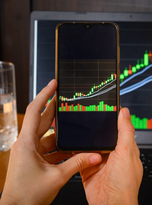 Manos sosteniendo un teléfono móvil que muestra un gráfico bursátil con velas verdes y rojas en tendencia alcista, frente a la pantalla de una computadora portátil.