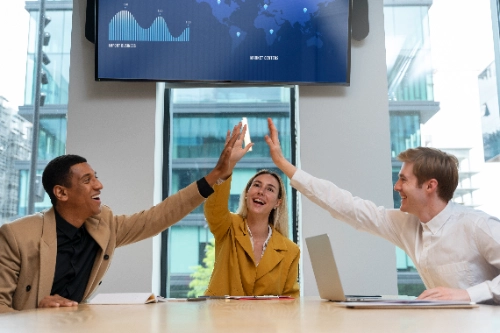 Tres colegas de negocios chocan los cinco alegremente celebrando un éxito en su oficina. Arriba de ellos se observa una pantalla grande que muestra gráficos globales de rendimiento financiero.
