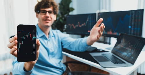 Un hombre sonriente con camisa azul muestra a la cámara un gráfico financiero en su celular, sentado en su escritorio de trabajo con múltiples monitores de fondo.