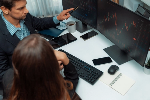 Vista desde arriba de un hombre y una mujer de negocios analizando gráficos bursátiles interactivos en dos monitores grandes en su escritorio de oficina.