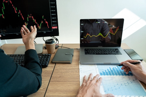 Dos personas en un escritorio analizan gráficos bursátiles financieros e informes impresos con una laptop y un monitor.