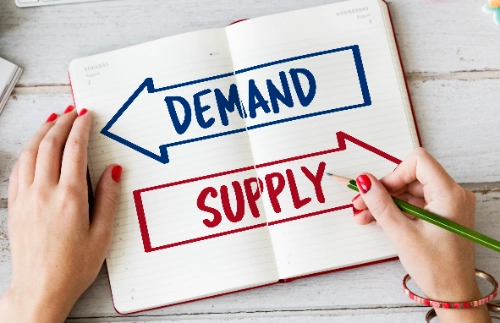 Un cuaderno abierto sobre una mesa de madera que muestra dos flechas dibujadas: una azul hacia la izquierda con la palabra "DEMAND" y otra roja hacia la derecha con la palabra "SUPPLY"