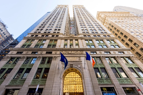 Toma en contrapicado de la imponente fachada del Equitable Building en Nueva York, destacando su gran entrada arqueada e iluminada con dos banderas ondeando arriba.