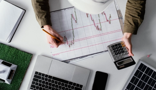 Vista aérea de una persona que escribe con un lápiz sobre un gráfico impreso mientras sostiene una calculadora en un escritorio con una computadora portátil y maquetas ecológicas.