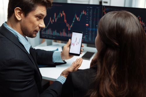 Un hombre de negocios le muestra un gráfico financiero en su teléfono móvil a su colega frente a dos monitores de oficina.