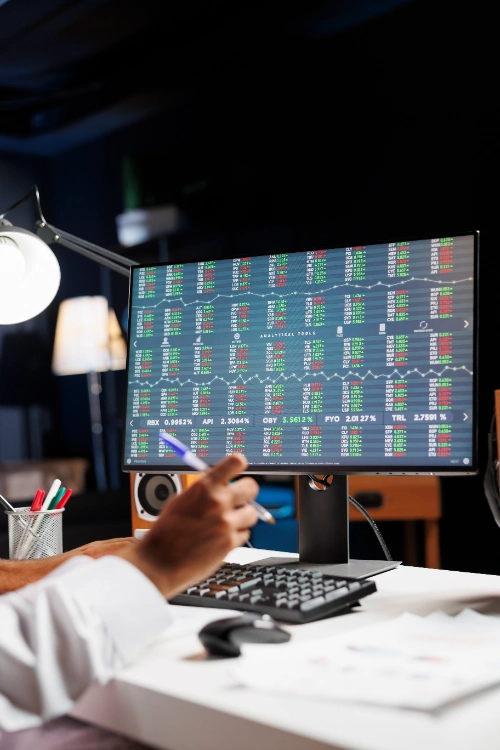 Una persona analiza datos en un monitor que muestra tablas y gráficos financieros detallados en verde y rojo, en un escritorio iluminado por una lámpara de noche.
