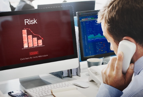 Un hombre habla por teléfono frente a una pantalla que muestra la palabra "Risk" y gráficos financieros descendientes.