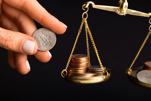 Una mano sostiene una moneda de un cuarto de dólar junto a una balanza dorada. Los platillos contienen montones de monedas sobre un fondo completamente negro.