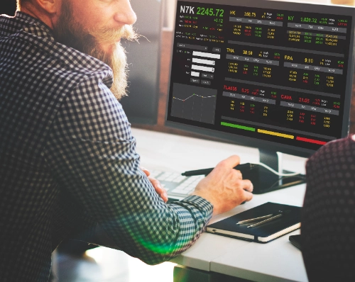 Un hombre con barba y camisa a cuadros analiza los mercados financieros en su escritorio. Frente a él se observa un monitor que muestra diferentes valores y gráficos de la bolsa.