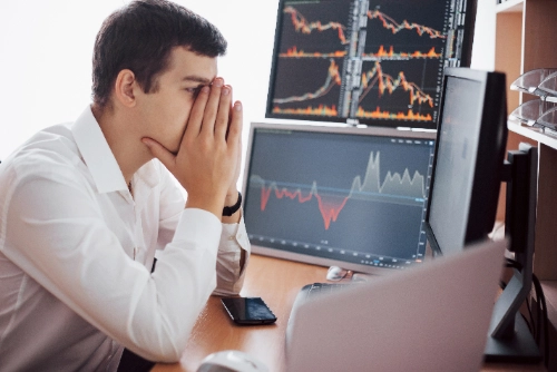 Un broker de camisa blanca se cubre el rostro frustrado frente a sus pantallas. Los monitores muestran gráficos de trading con marcadas pérdidas financieras.