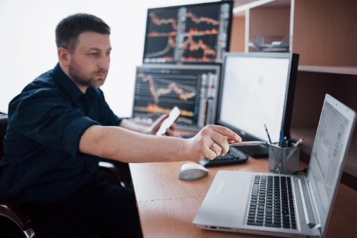 Un analista financiero trabaja en su escritorio rodeado de múltiples pantallas de trading. Con una mano apunta con un bolígrafo hacia su laptop mientras sostiene su teléfono con la otra.