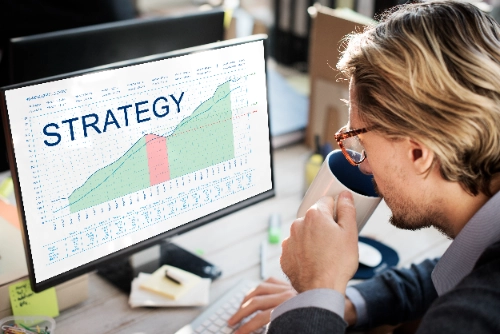 Vista lateral de un hombre rubio con gafas bebiendo de una taza blanca mientras observa un monitor que muestra un gráfico lineal ascendente con la palabra "STRATEGY" en letras grandes.