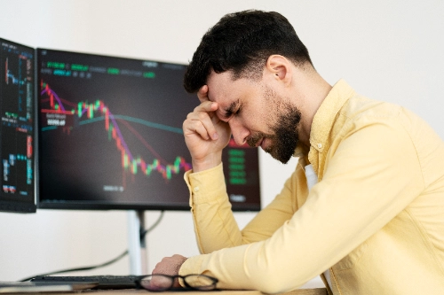 Un hombre con camisa amarilla se toca la frente frustrado por las pérdidas financieras de su negocio. Al fondo se observan las pantallas de su computadora mostrando caídas drásticas en los gráficos de trading.