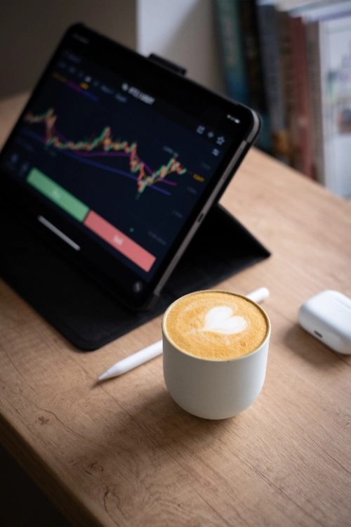 Una taza blanca con café latte art de corazón sobre un escritorio de madera, junto a un lápiz digital, unos auriculares inalámbricos y una tableta inclinada que muestra gráficos financieros de trading.