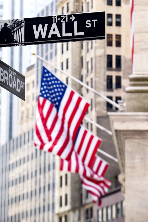 Un letrero de la calle "WALL ST" en primer plano con banderas de los Estados Unidos y edificios corporativos difuminados al fondo.
