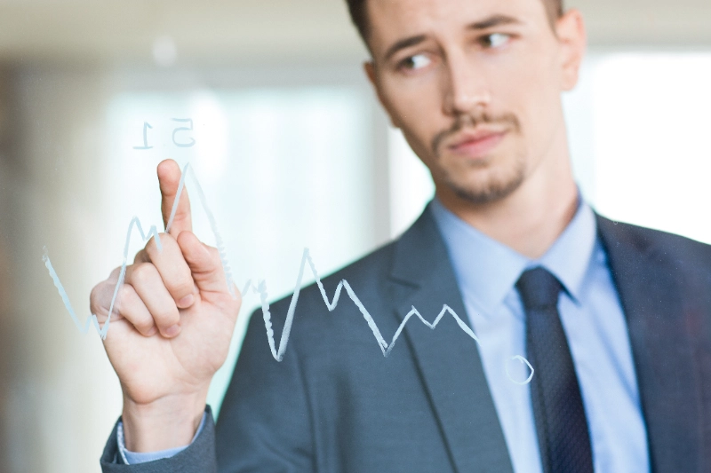Empresario trajeado señalando el punto más alto de un gráfico de líneas trazado sobre un panel transparente.