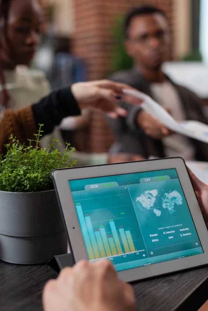 Plano en primer plano de una tableta digital mostrando estadísticas de marketing global y gráficos de barras, con un equipo de trabajo intercambiando documentos en segundo plano.