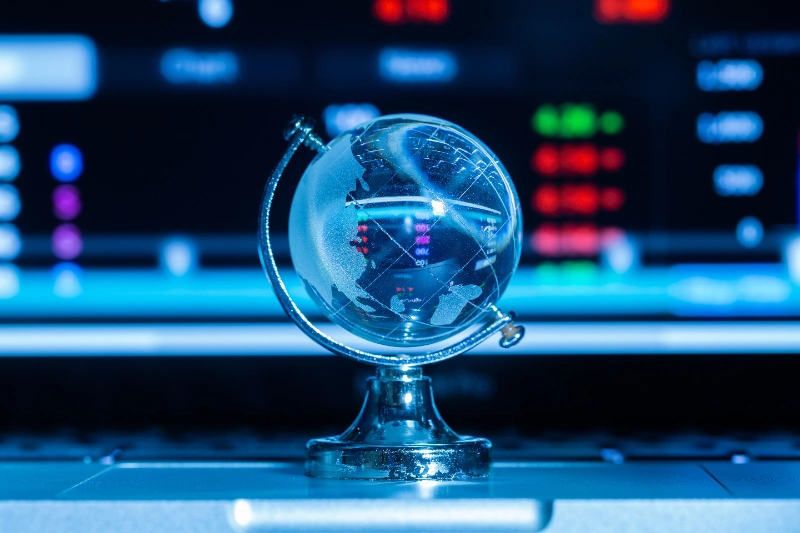 Globo terráqueo de cristal sobre un portátil con una pantalla de fondo que muestra datos y gráficos bursátiles reflejados.