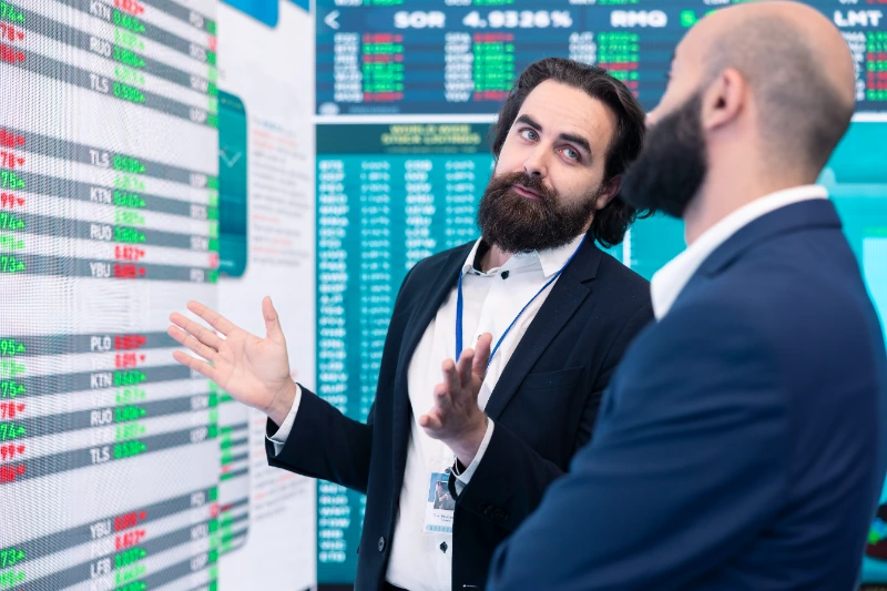 Analista financiero gesticulando mientras explica datos e indicadores de mercado en una gran pantalla bursátil a su colega.