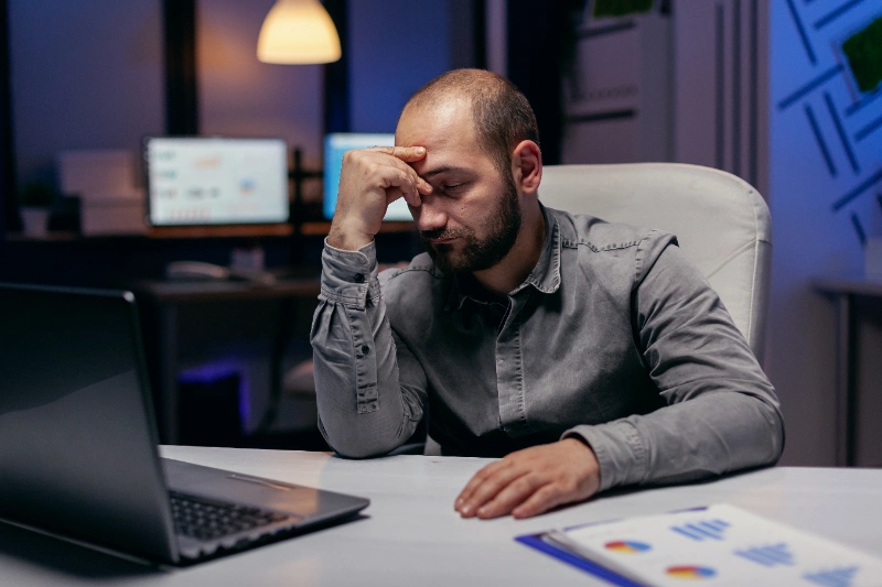 Ejecutivo fatigado reclinando la cabeza sobre su mano frente a una computadora portátil durante una jornada nocturna de trabajo en la oficina.