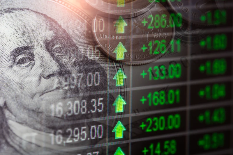 Billete de dólar y monedas superpuestos con gráficos bursátiles e indicadores verdes de tendencia al alza.